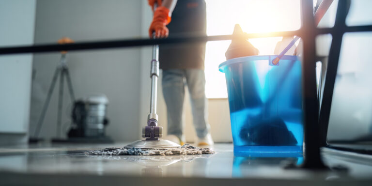 A cleaning woman is standing inside a building holding with a blue tank on the side facilities for tidying up in her hand